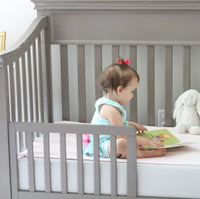 Photo of a child in a toddler bed, reading a picture book with two plus rabbits looking on.  The ...