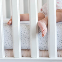 This image shows a child sleeping in a white crib, with one foot pushing through the slat.  The c...