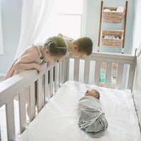 Photo of a very young baby in a crib with two older children leaning over the edge and gazing dow...