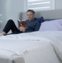 Photo of a comfortable bed in a bright room with neutral upholstered head and foot boards. A man ...