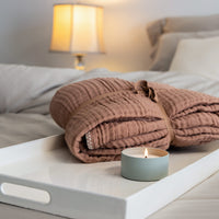 Photo of a terracotta cotton muslin throw folded on a tray with a lit candle.  The tray is on a b...